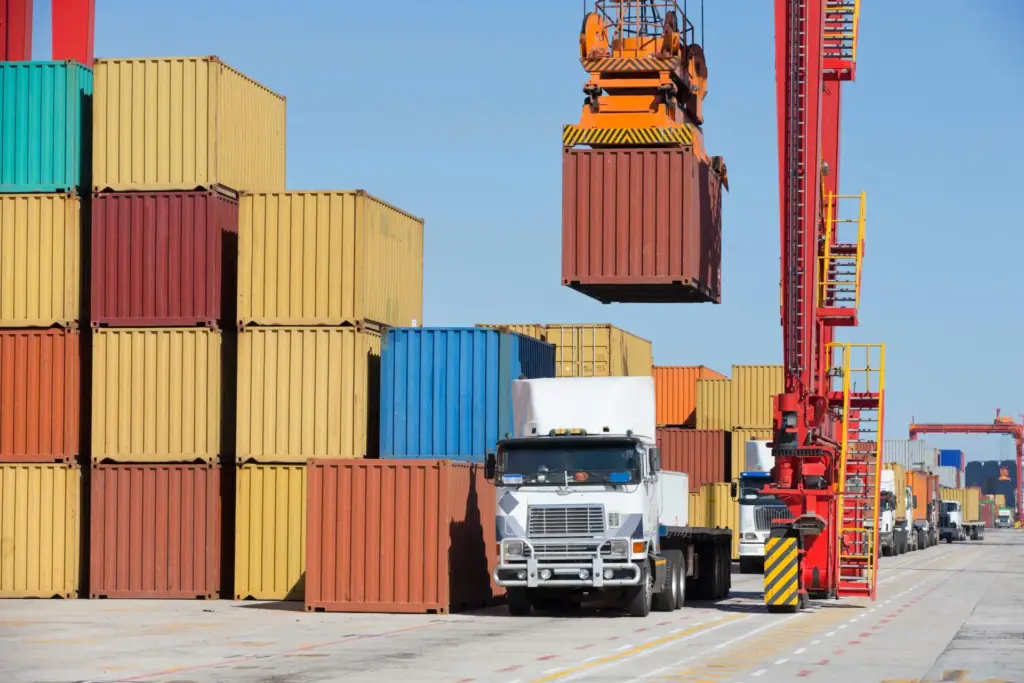 Containerkran hebt einen Container über gestapelte Container, davor ein LKW